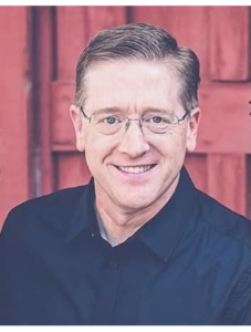 Image of Pastor Travis in blue shirt smiling in front of red background.